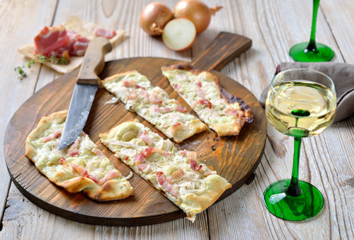 In Stücke geschnittener Flammkuchen mit Speck und Zwiebeln auf einem Holzbrett, daneben ein Glas Weißwein