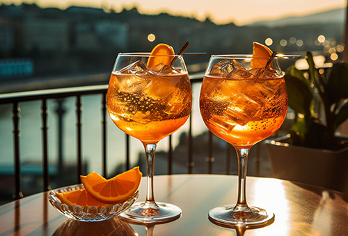 Zwei Gläser Aperol Spritz auf einem Tisch auf einem Balkon mit Blick über einen Fluss im Sonnenuntergang