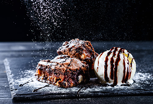 Zwei Stücke Brownie, daneben eine Kugel Vanilleeis, über allem Schokosoße und Puderzucker