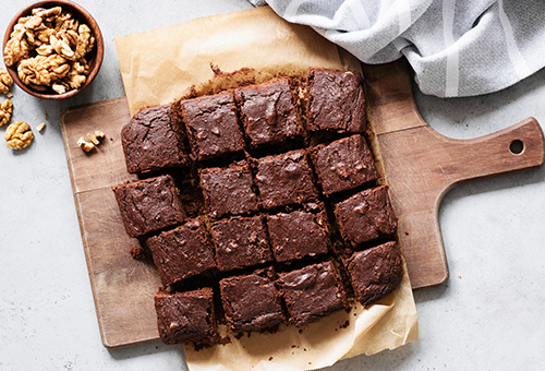 Brownie in quadratische Stücke geschnitten auf einem Holzbrett, daneben ein Schälchen mit Walnüssen