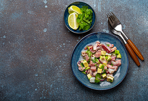 Ceviche mit Avocado in einer blauen Schale, in einer kleinen Schale daneben Koriander und Limetten