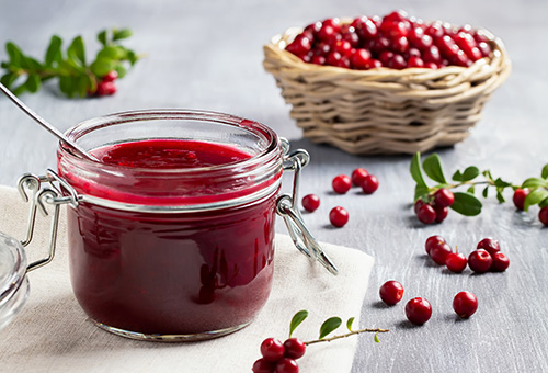 Marmelade aus Preiselbeeren im Glas im Vordergrund, im Hintergrund ein Körbchen mit reifen Preiselbeeren