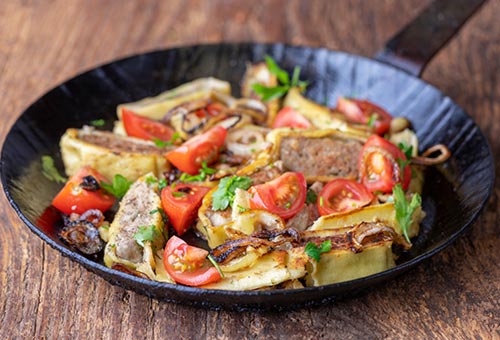 Maultaschenscheiben angebraten mit kleinen Tomaten und Zwiebeln in einer Gusspfanne garniert mit frischen Kräutern