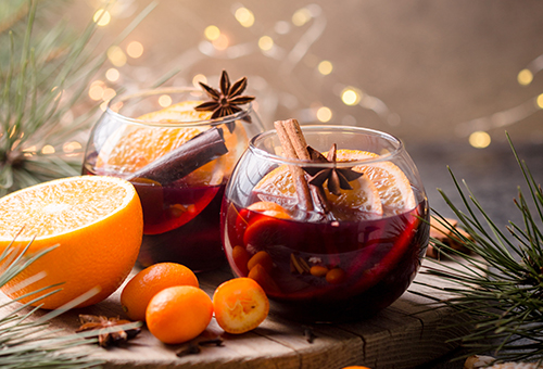 Zimtlikör in runden Gläsern angerichtet auf einem Holzbrett weihnachtlich dekoriert mit Orangen, Lichterkette und Zimtstangen.